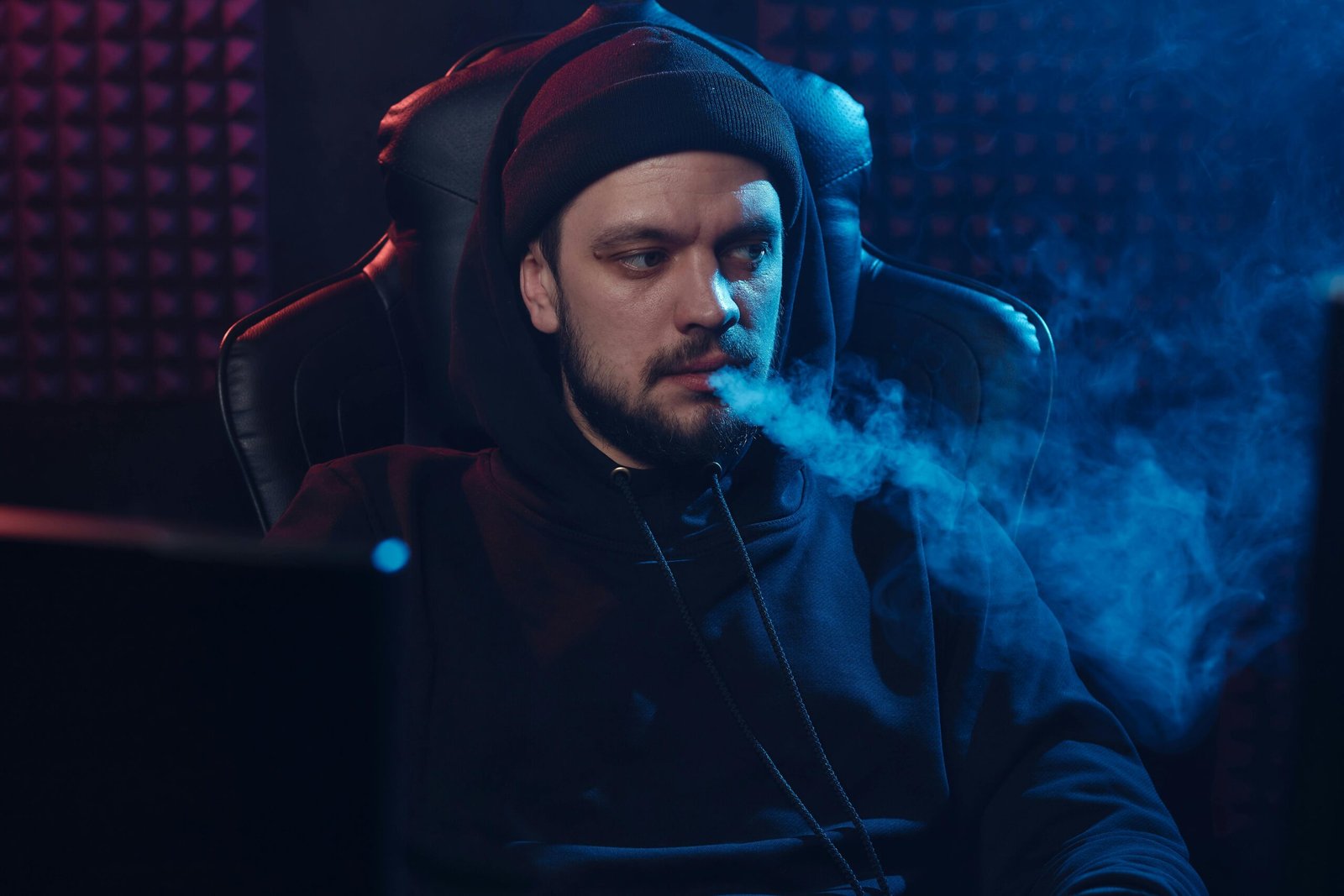 Bearded man in dark room exhaling smoke, wearing hoodie and beanie, depicted in moody lighting.